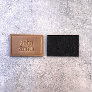 以下が含まれることがあります： 「John Smith」のエンボス加工が施された長方形の薄茶色の革パッチと、黒い布パッチ。 革パッチには隆起した縁があり、テキストは中央に配置されています。