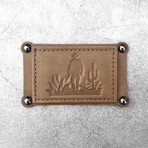 Pode incluir: Um patch retangular de couro castanho claro com uma borda costurada e cantos arredondados, preso por quatro rebites prateados. O patch apresenta um desenho em relevo de um cowboy com chapéu, cactos e plantas do deserto.