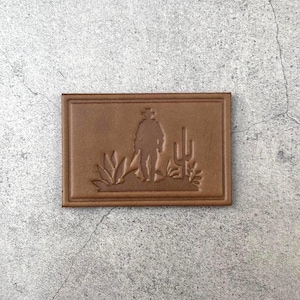 以下が含まれることがあります： カウボーイのシルエットがサボテンなどの植物がある砂漠の風景に立っている、長方形の茶色の革パッチ。 パッチには隆起した境界線があります。