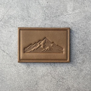 May include: A rectangular, brown leather patch featuring a mountain range design. The mountains are embossed within a raised border, creating a textured effect. The patch is a warm, earthy tone, suitable for various applications.