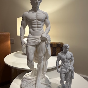 Puede incluir: Dos estatuas blancas de una figura masculina musculosa, una grande y otra pequeña, están sobre mesas redondas blancas. La estatua más grande mide aproximadamente 76 cm de altura, y la estatua más pequeña mide unos 30 cm de altura. Ambas figuras están cubiertas con tela.