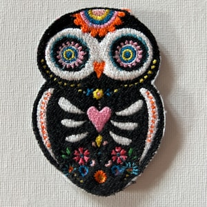 Könnte beinhalten: Gestickter Eulenaufnäher im Stil des Día de los Muertos. Die Eule ist schwarz mit weißen, skelettartigen Markierungen, einem rosa Herzen und farbenfrohen floralen und geometrischen Details. Die Augen der Eule sind groß und detailliert mit blauen und gelben Akzenten.