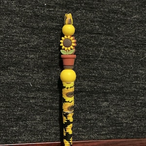 Puede incluir: Bolígrafo decorativo con temática de girasoles. El bolígrafo tiene un barril negro adornado con girasoles amarillos. La parte superior del bolígrafo tiene cuentas amarillas, un girasol y una maceta pequeña. El bolígrafo mide unos 15 cm de largo.