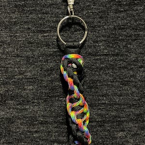 Puede incluir: Un llavero trenzado negro y arcoíris con un mosquetón y un anillo plateados. El llavero presenta un diseño tejido con hilos negros y de colores vibrantes del arcoíris, creando un accesorio visualmente atractivo.