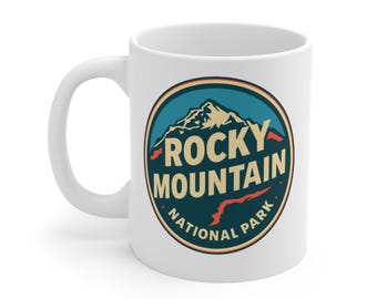 May include: White ceramic mug featuring a circular design with the text "ROCKY MOUNTAIN NATIONAL PARK". The design includes a mountain range in beige and a blue background with red and orange accents. The mug has a curved handle.