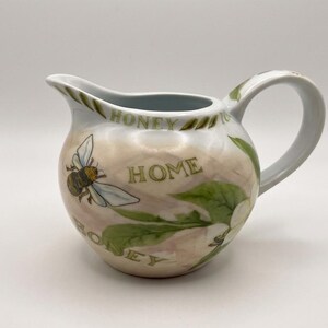 May include: A white ceramic creamer pitcher featuring a painted bee, green leaves, and the text "HONEY" and "HOME" in gold lettering. The pitcher has a rounded body, a curved handle, and a pouring spout with a smooth, glossy finish.