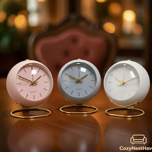 May include: Three spherical quartz clocks in pastel colors: pink, gray, and white. Each clock has a gold-colored base and hands. The clock faces display simple, minimalist designs. The clocks are arranged on a dark wooden surface.