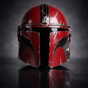 May include: A red and black Mandalorian helmet with battle-worn details. The helmet features a rounded top with a central stripe and angular visor. The surface has a distressed finish, suggesting use in combat. The helmet is displayed on a reflective surface.