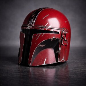 May include: A red and black Mandalorian helmet with silver scratches and wear marks. The helmet has a distinctive T-shaped visor and a rounded top. The helmet is a prop or collectible item.