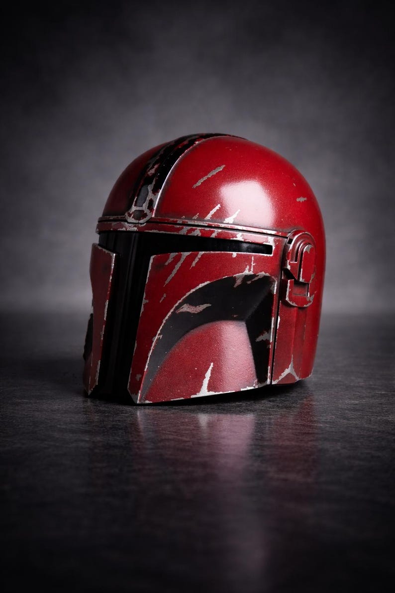 May include: A red and black Mandalorian helmet with silver scratches and wear marks. The helmet has a distinctive T-shaped visor and a rounded top. The helmet is a prop or collectible item.