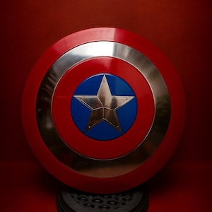 May include: A red, white, and blue shield with a silver ring and a gold star in the center. The shield is round and rests on a black base, set against a red background.