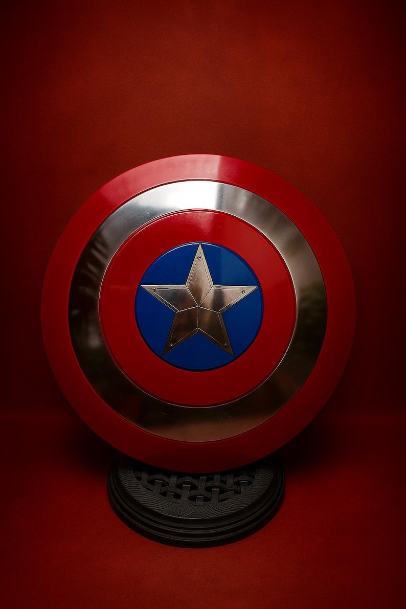 May include: A red, white, and blue shield with a silver ring and a gold star in the center. The shield is round and rests on a black base, set against a red background.