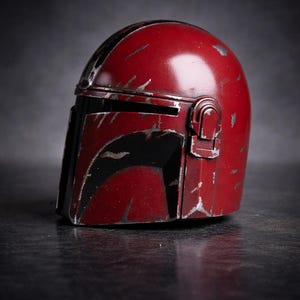 May include: A weathered, red Mandalorian helmet with black accents. The helmet has visible scratches and wear, suggesting a battle-worn appearance. The design includes a visor and a side attachment, typical of the iconic helmet.
