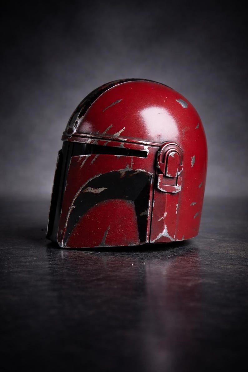 May include: A weathered, red Mandalorian helmet with black accents. The helmet has visible scratches and wear, suggesting a battle-worn appearance. The design includes a visor and a side attachment, typical of the iconic helmet.