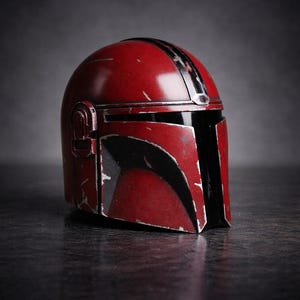 May include: A red Mandalorian helmet with black accents and a weathered appearance. The helmet has a visor and a T-shaped opening for the eyes. The helmet is a prop from the Star Wars franchise.