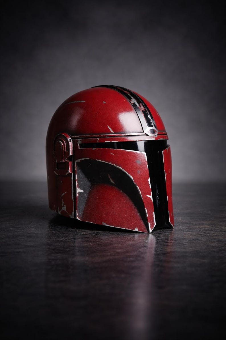 May include: A red Mandalorian helmet with black accents and a weathered appearance. The helmet has a visor and a T-shaped opening for the eyes. The helmet is a prop from the Star Wars franchise.