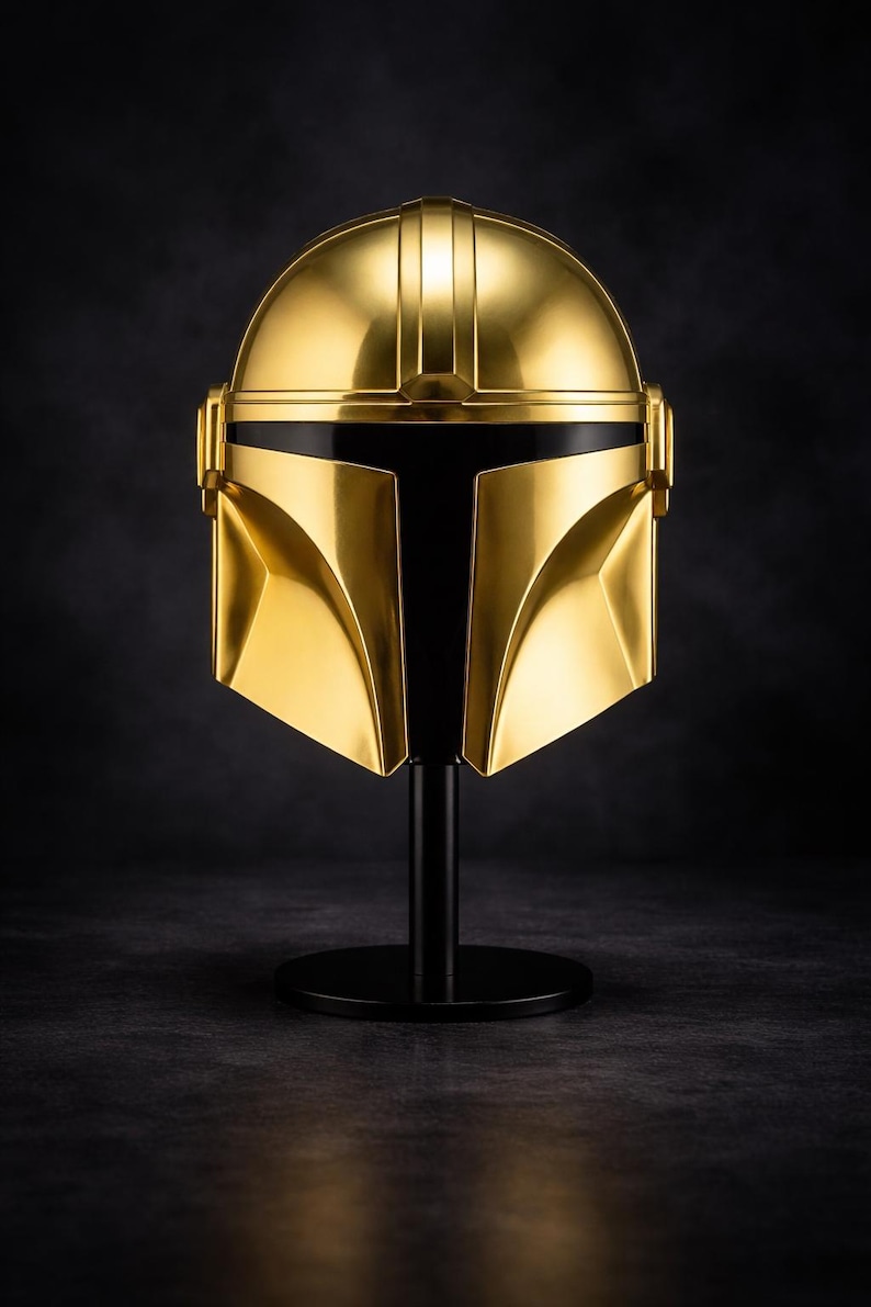 May include: A gold-colored helmet with a black visor, displayed on a black stand. The helmet has a smooth, reflective surface and a distinctive T-shaped visor. The helmet is a replica of a Mandalorian helmet.