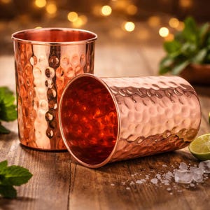 Könnte beinhalten: Zwei Kupfer-Moscow-Mule-Becher mit gehämmerten Texturen. Ein Becher steht aufrecht, der andere liegt auf der Seite. Die Becher stehen auf einer Holzoberfläche mit verschwommenen Hintergrundlichtern, Minzblättern und Limettenscheiben.