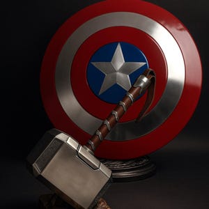 May include: A silver and brown hammer with a leather strap handle rests on a wooden base, in front of a red, white, and blue shield with a star emblem. The items are displayed against a dark background.