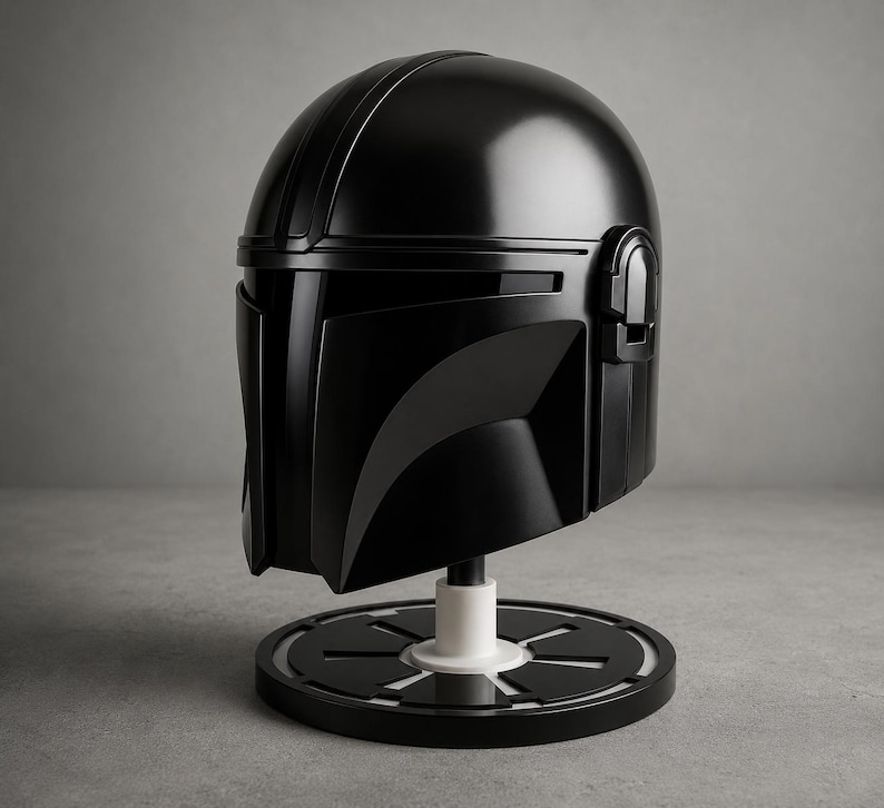 Puede incluir: Una r&eacute;plica de casco negro mandaloriano, exhibida sobre un soporte circular en blanco y negro. El casco tiene un acabado brillante con elementos de dise&ntilde;o detallados. El soporte tiene un soporte cil&iacute;ndrico blanco.