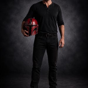 May include: A person wearing a black long-sleeve shirt and black jeans holds a red and black helmet. The helmet has a T-shaped visor and is likely a prop or costume piece. The person is wearing brown boots.