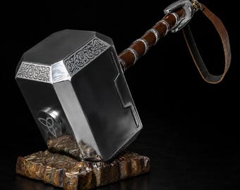 May include: A silver-colored hammer with a rectangular head featuring intricate knotwork engravings and a circular triquetra symbol. The handle is brown with silver accents and a leather strap, resting on a textured, bronze-colored rock base.