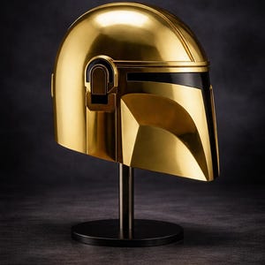 May include: A gold-colored Mandalorian helmet, displayed on a black stand. The helmet has a sleek, reflective surface with black accents. The design is angular and futuristic, with a visor and distinctive shape.