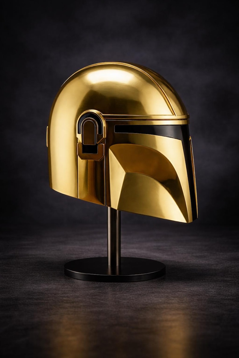 May include: A gold-colored Mandalorian helmet, displayed on a black stand. The helmet has a sleek, reflective surface with black accents. The design is angular and futuristic, with a visor and distinctive shape.