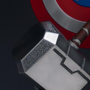 May include: A close-up of Thor's hammer, Mjolnir, with a silver head and a brown leather handle. The hammer has intricate engraved designs and is set against a backdrop of Captain America's shield.