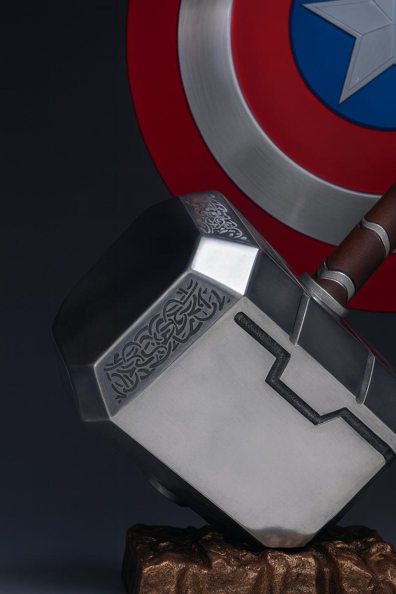 May include: A close-up of Thor's hammer, Mjolnir, with a silver head and a brown leather handle. The hammer has intricate engraved designs and is set against a backdrop of Captain America's shield.