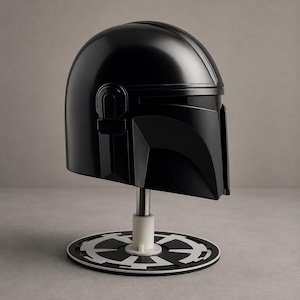 Puede incluir: Un casco negro mandaloriano, exhibido en un soporte con una base circular blanca y negra. El casco tiene un acabado liso y brillante y es una r&eacute;plica del ic&oacute;nico casco de la serie Star Wars.