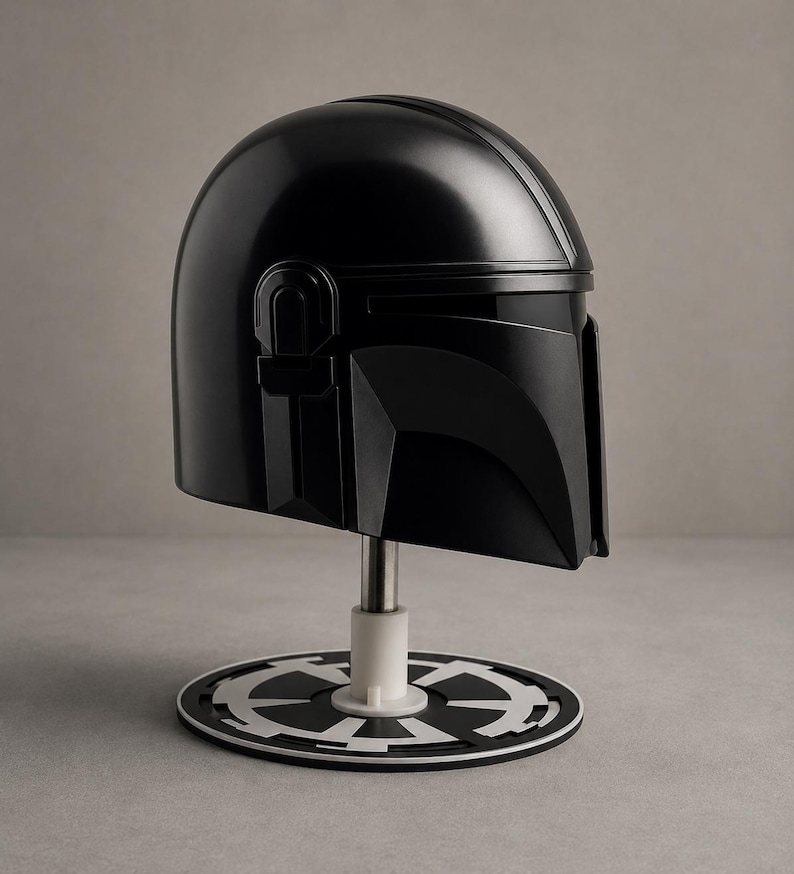 Puede incluir: Un casco negro mandaloriano, exhibido en un soporte con una base circular blanca y negra. El casco tiene un acabado liso y brillante y es una r&eacute;plica del ic&oacute;nico casco de la serie Star Wars.
