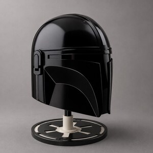 Puede incluir: Un casco negro brillante de Mandaloriano, exhibido en un soporte con una base circular blanca y negra. El casco presenta un dise&ntilde;o elegante y angular con una visera y detalles distintivos.