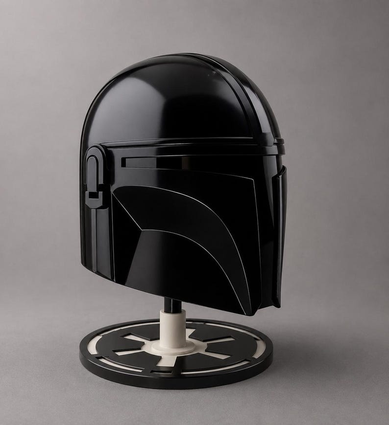 Puede incluir: Un casco negro brillante de Mandaloriano, exhibido en un soporte con una base circular blanca y negra. El casco presenta un dise&ntilde;o elegante y angular con una visera y detalles distintivos.