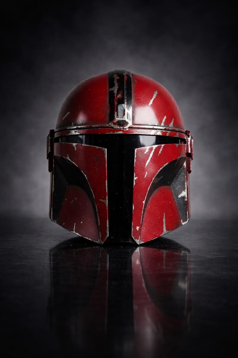 May include: A red and black Mandalorian helmet with battle-worn details. The helmet features a rounded top with a central stripe and angular visor. The surface has a distressed finish, suggesting use in combat. The helmet is displayed on a reflective surface.