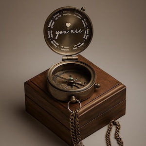 May include: A vintage compass with the words "you are" and a heart symbol engraved on the lid. The compass is set in a wooden box with a brass chain. The compass needle points north.