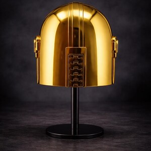 May include: A gold-colored helmet with a glossy finish, displayed on a black stand. The helmet has vertical lines on top and a rectangular panel with a series of horizontal bars. The stand has a circular base.