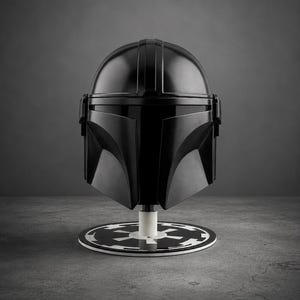 Puede incluir: Un casco negro de Mandaloriano, exhibido sobre un soporte circular blanco y negro. El casco tiene un acabado brillante y est&aacute; montado sobre un poste blanco. El soporte tiene un di&aacute;metro de aproximadamente 20 cm.