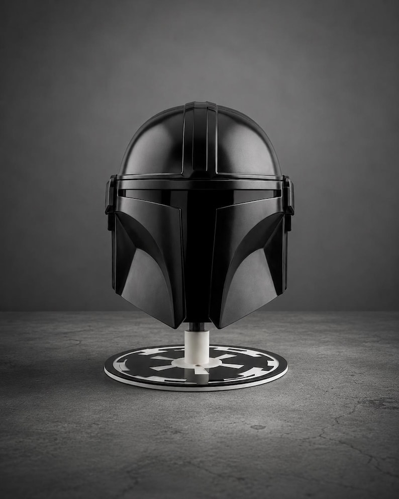Puede incluir: Un casco negro de Mandaloriano, exhibido sobre un soporte circular blanco y negro. El casco tiene un acabado brillante y est&aacute; montado sobre un poste blanco. El soporte tiene un di&aacute;metro de aproximadamente 20 cm.