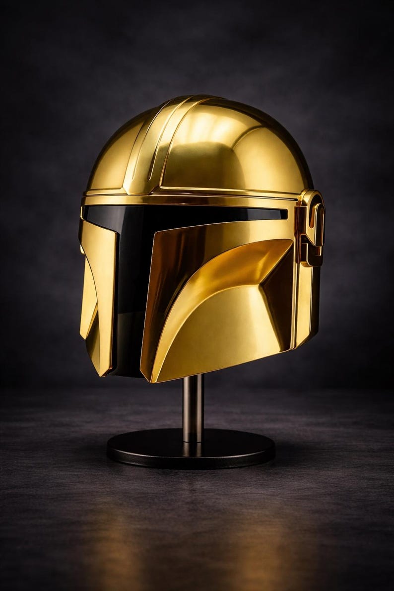 May include: A gold-colored Mandalorian helmet, displayed on a black stand. The helmet has a sleek, metallic finish with a black visor. The design features angular shapes and a prominent T-shaped visor.