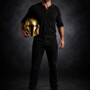 May include: A full-length shot of a person holding a gold-colored Mandalorian helmet. The person is wearing a black long-sleeve shirt, black jeans, and brown boots. The background is a dark gray.