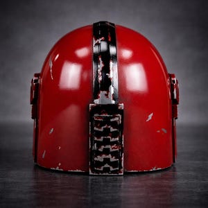 May include: A red helmet with a distressed finish, featuring a black stripe down the center and rectangular details on the back. The helmet has a glossy appearance with visible wear and tear.