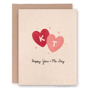 May include: A greeting card with a light beige background and a brown envelope. The card features two heart shapes, one red with a white "K" and one pink with a white "T". The text "Happy You + Me Day" is written below the hearts.