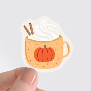 May include: A white ceramic mug with a pumpkin design and a brown handle. The mug is filled with a white whipped cream topping and two cinnamon sticks.