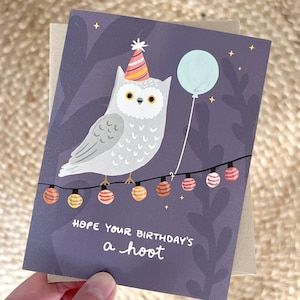 May include: A birthday greeting card featuring a cartoon owl wearing a party hat and holding a balloon. The owl is perched on a string of colorful lights against a dark purple background. The text reads, "Hope your birthday's a hoot."