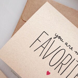 May include: A card with the text "you are my favorite" in black script font with a small red heart below the text. The card is on a brown paper background.