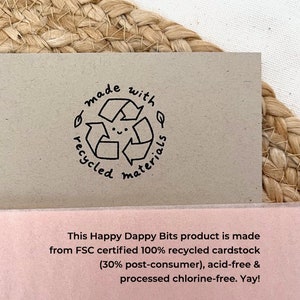 May include: A close-up of a brown card with a black and white stamp that says "made with recycled materials". The stamp features a smiling recycling symbol. The card also has text that says "This Happy Dappy Bits product is made from FSC certified 100% recycled cardstock (30% post-consumer), acid-free & processed chlorine-free. Yay!"