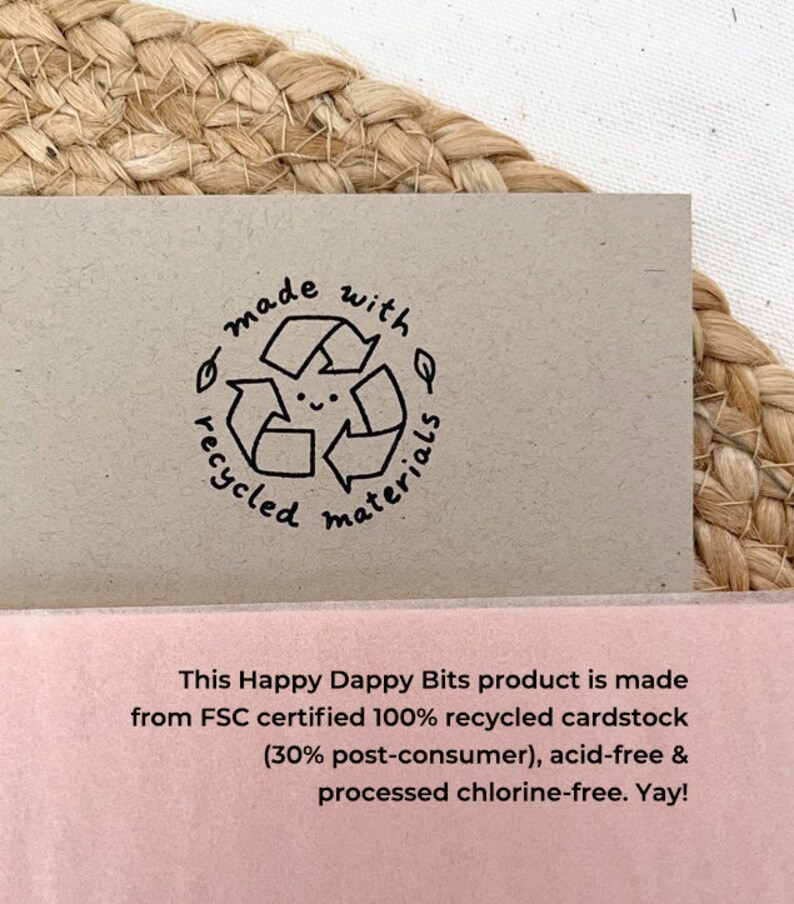 May include: A close-up of a brown card with a black and white stamp that says "made with recycled materials". The stamp features a smiling recycling symbol. The card also has text that says "This Happy Dappy Bits product is made from FSC certified 100% recycled cardstock (30% post-consumer), acid-free & processed chlorine-free. Yay!"