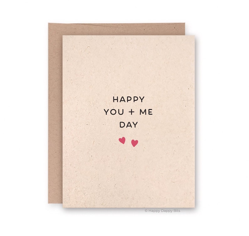 May include: A greeting card with a beige front and a brown envelope. The front of the card has the words "HAPPY YOU + ME DAY" in black, with two small pink hearts below the text. The card is a simple, minimalist design.
