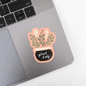 May include: A pink sticker with a black pot and green leaves. The pot has gold glitter and the text "plant lady" written in white.